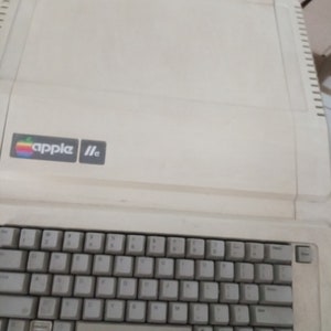 May include: A vintage white Apple IIc computer with a keyboard and the Apple logo on the front.