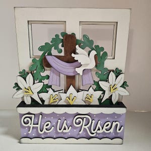 Puede incluir: Un letrero de Pascua de madera con un marco de ventana blanco, una cruz morada y marrón, una paloma blanca, hojas verdes, flores blancas y una maceta morada con el texto "He is Risen".
