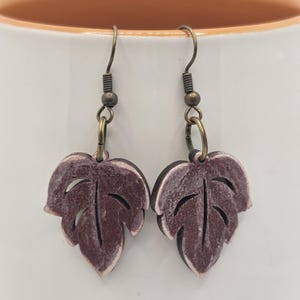 May include: A pair of dangle earrings featuring a leaf design. The leaves are made of wood and painted a deep purple color. The earrings have a bronze-colored hook.