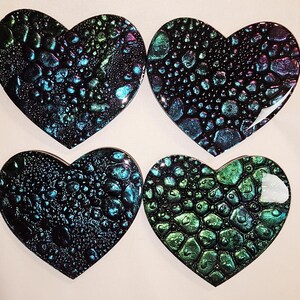 Pode incluir: Quatro  tapetes de copo em forma de coração feitos de resina com acabamento preto, verde e iridescente. Os tapetes têm uma superfície texturizada que se assemelha a escamas ou bolhas.