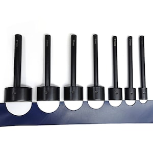 Puede incluir: Conjunto de 7 punzones de cuero negros de diferentes tamaños: 4,5 mm, 4 mm, 3,5 mm, 3 mm, 2,5 mm, 2 mm y 2,1 mm. Los punzones están dispuestos en orden de tamaño, con el punzón más grande a la izquierda y el más pequeño a la derecha. Los punzones están sobre una superficie azul.