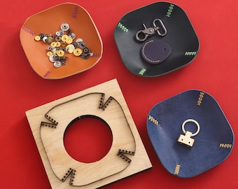 Op de afbeelding: Vier lederen ambachtelijke projecten, waaronder een ronde lederen pons, een klein lederen dienblad met metalen studs, een blauw lederen dienblad met een sleutelhanger en een bruin lederen dienblad met metalen klinknagels en decoratieve stiksels.