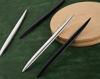 May include: Three thin, sharp tools, two silver and one black, are arranged on a green surface. One silver tool is on a wooden surface.