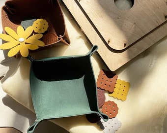 Op de afbeelding: Handgemaakte leren trays in bruin en groen, met decoratieve leren uitsparingen in de vorm van bloemen en koekjes. Een houten bord met een cirkelvormig gat is ook zichtbaar. Geschikt voor kleine items.