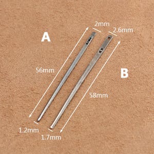 May include: Two silver leather sewing needles, labeled A and B, with measurements in millimeters. Needle A is 56 mm long and 1.2 mm wide. Needle B is 58 mm long and 1.7 mm wide. The needles have a small eye at one end and a pointed tip at the other.