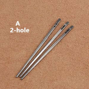 May include: Three silver leather sewing awls with two holes each. The awls are laid out on a brown surface. The text "A 2-hole" is visible in the image.