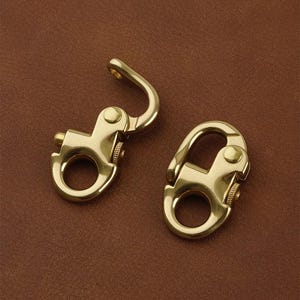 May include: Two brass-colored swivel snap hooks. One hook is open, revealing the spring-loaded mechanism. The other hook is closed. The hooks have a circular ring at the base and a curved hook at the top. The background is a solid brown color.