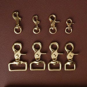 May include: A set of ten brass swivel clasps, some with a D-ring, in various sizes. The clasps are arranged in two rows on a brown leather surface.