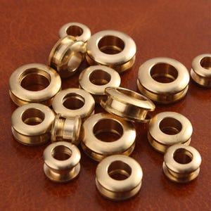 May include: A collection of gold-toned metal eyelets of various sizes. The eyelets are arranged in a random pattern on a brown leather surface.