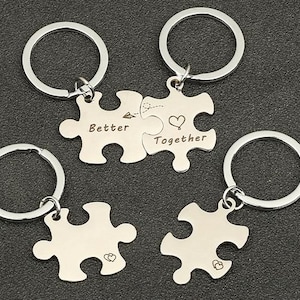 Può includere: Due portachiavi argentati con pezzi di puzzle che si incastrano. I pezzi sono incisi con le parole "Better" e "Together".