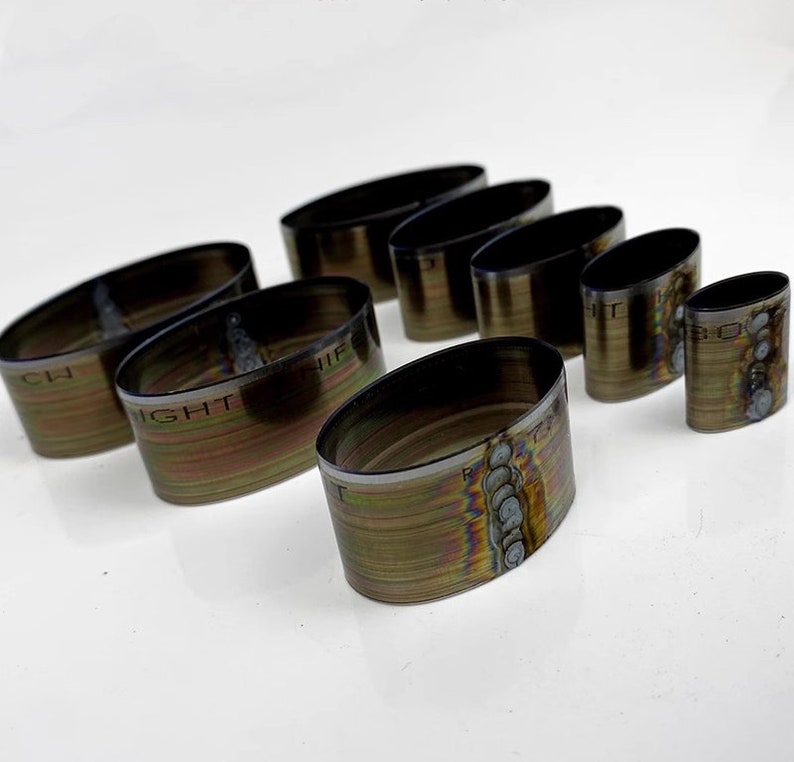 Op de afbeelding: Set van acht ovale metalen ringen met iriserende regenboogkleuren. De ringen hebben verschillende maten en zijn gelabeld met tekst, waaronder "RIGHT", "CH" en "NIF".