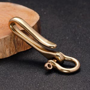 May include: A solid brass key clip with a curved design. The clip features a hook on one end and a circular loop on the other, connected by a swivel. The brass has a polished finish, reflecting light. The key clip is shown on a dark surface.