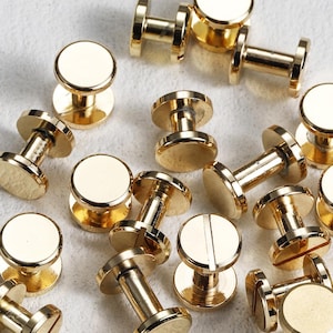 May include: A collection of gold-colored metal Chicago screws, also known as binding posts or screw posts. Each piece features a smooth, flat circular head and a cylindrical shaft with a threaded screw mechanism for fastening materials together.