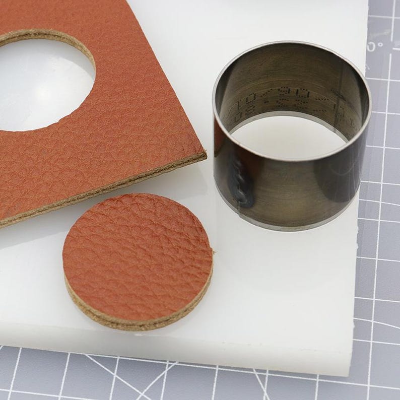May include: A brown leather circle cut out with a metal circle cutter on a white cutting mat. The cutter has a measurement of 1 1/2 inches.