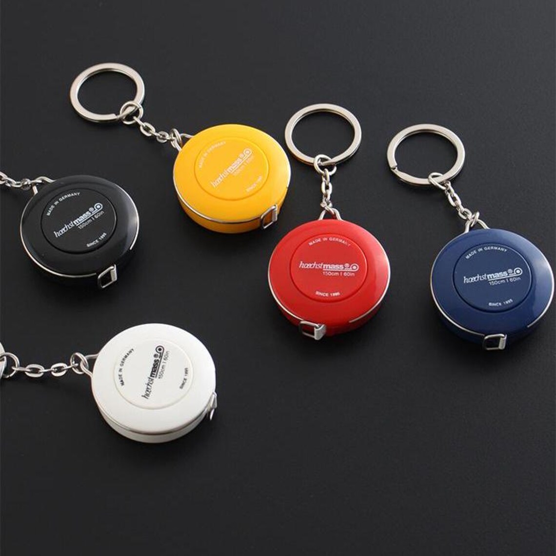 May include: Five colourful keychain tape measures with a black, white, yellow, red, and blue design. Each tape measure has the text "hochstmass 1.5" printed on it.