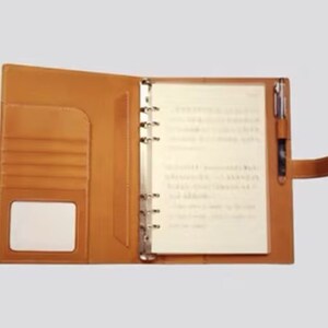 Könnte beinhalten: Ein braunes, ledergebundenes Notizbuch mit einem Stifthalter und einer durchsichtigen Plastiktasche für einen Notizblock. Das Notizbuch hat eine Tasche auf der Vorderseite und ein transparentes Plastikfenster.