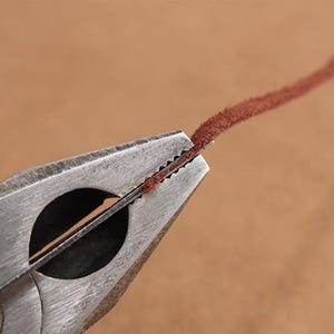 May include: Close-up of a metal tool with a serrated edge used to crimp leather cord. The cord is brown and is being held in place by the tool.