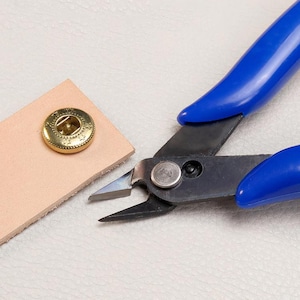 May include: Close-up of a leathercraft project with a gold snap and blue-handled pliers. The pliers are positioned to cut the leather, with the snap already attached to a piece of light brown leather.