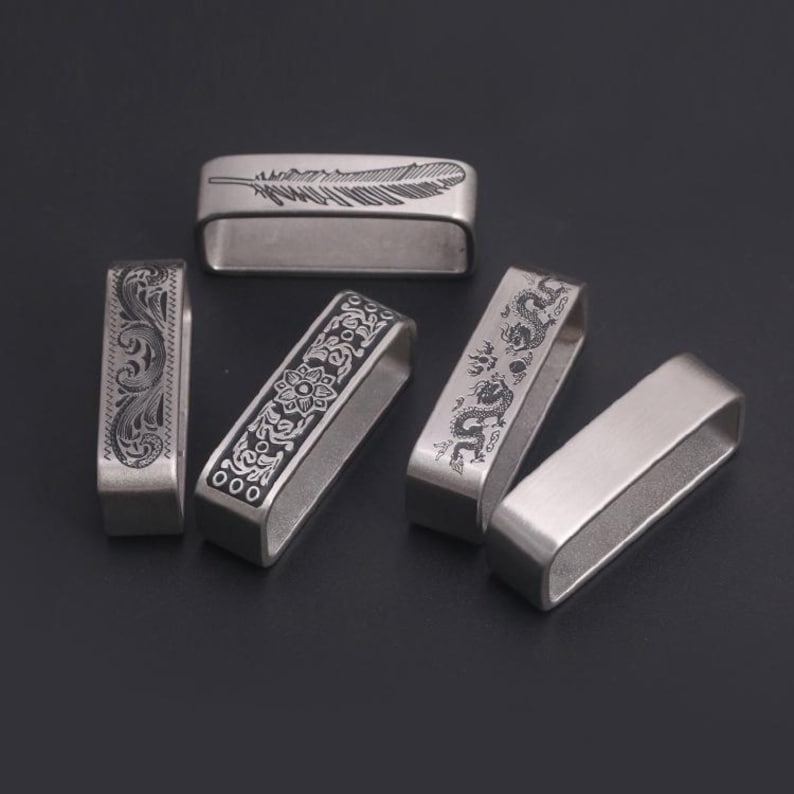 May include: Five silver metal belt buckle keepers with different engraved designs. The designs include a feather, a floral pattern, a dragon, and two plain keepers.