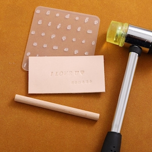 May include: A leather stamping kit with a clear alphabet stamp, a wooden mallet, and a wooden handle. The leather is stamped with the words "I LOVE U" and a heart.