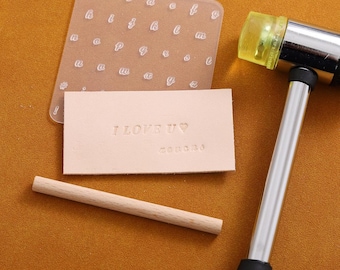 Könnte beinhalten: Ein Lederprägesatz mit einem transparenten Alphabet-Stempel, einem Holzhammer und einem Holzgriff. Das Leder ist mit den Worten "I LOVE U" und einem Herzen geprägt.