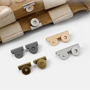 Puede incluir: Surtido de cierres magnéticos en varios acabados metálicos, incluyendo plata, oro y bronce. Los cierres se muestran con una tela tejida, lo que sugiere su uso en bolsos u otros accesorios. Los cierres son redondos y rectangulares.