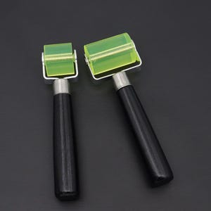 Puede incluir: Dos herramientas de rodillo verdes y negras con mangos de madera. Los rodillos están hechos de un material transparente y flexible y se utilizan para alisar las superficies.