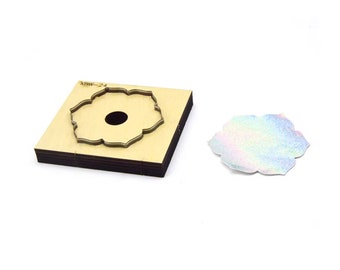 Puede incluir: Una herramienta de troquelado de madera con un diseño en forma de flor. El troquel se utiliza para cortar formas de papel, tela u otros materiales. El troquel se muestra junto a una muestra de la forma cortada, que está hecha de material iridiscente.