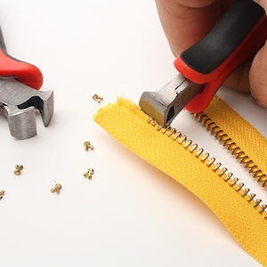 May include: A pair of red pliers with black handles are used to remove the metal teeth from a yellow zipper. The teeth are scattered around the zipper.