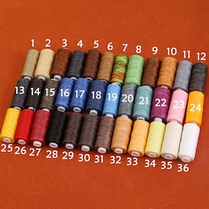 Pode incluir: Um conjunto de 36 carretéis de fio encerado em várias cores, numerados de 1 a 36. Os carretéis estão dispostos em duas filas, com os números visíveis na fila superior.