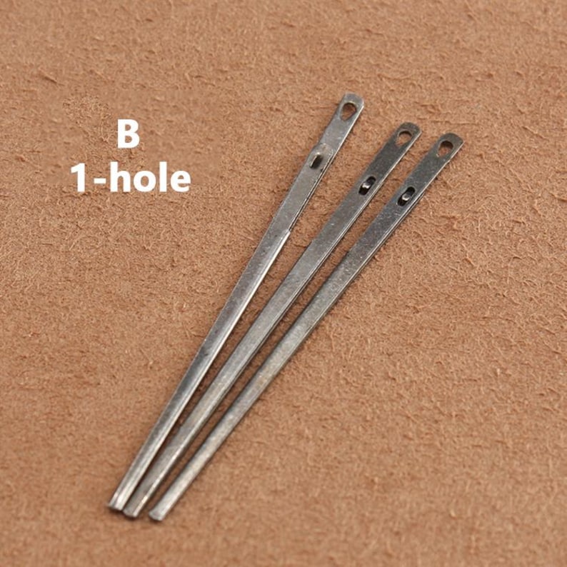 May include: Three silver leather sewing awls with a single hole at the top. The awls are laid out on a brown suede surface. The text "B 1-hole" is visible in the top left corner of the image.