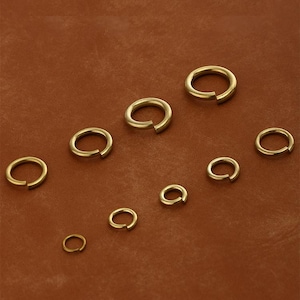 Op de afbeelding: Een verzameling goudkleurige splitringen van verschillende maten, gerangschikt op een bruin oppervlak. De ringen zijn open, met een kleine opening aan één kant. Het metaal heeft een glanzende afwerking, die het licht reflecteert.