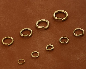 Può includere: Una collezione di anelli aperti dorati di varie dimensioni, disposti su una superficie marrone. Gli anelli sono aperti, con una piccola fessura su un lato. Il metallo ha una finitura lucida, che riflette la luce.