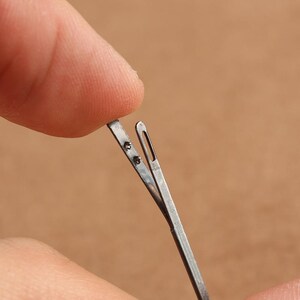 May include: A close-up of a silver threading tool with a small loop at the end. The tool is held open by a finger.