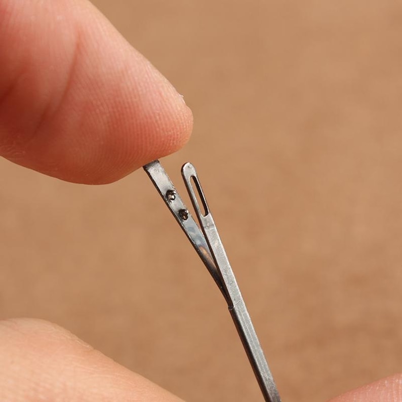 May include: A close-up of a silver threading tool with a small loop at the end. The tool is held open by a finger.