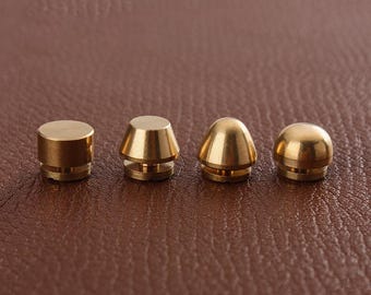 May include: Four gold-colored metal caps with different shapes. The caps are arranged in a row on a brown leather surface.