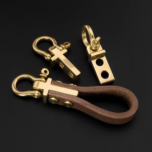 May include: Three brass keychains with a brown leather loop. The keychains have a cross-shaped design and a sturdy clasp. The leather loop is attached with brass rivets. The background is black.
