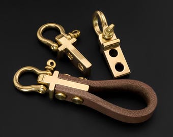 May include: Three brass keychains with a brown leather loop. The keychains have a cross-shaped design and a sturdy clasp. The leather loop is attached with brass rivets. The background is black.