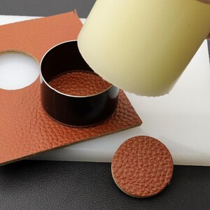 May include: A brown leather circle is being cut out using a round leather punch tool. The tool is being pressed down on the leather with a white cylinder. A second brown leather circle has already been cut out and is sitting on a white surface.