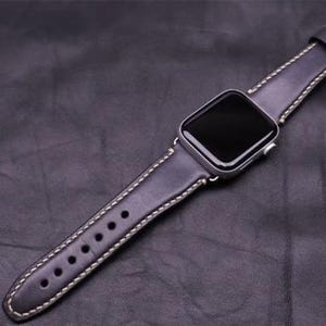 Op de afbeelding: Een donkerpaarse leren horlogeband met witte stiksels. De band is ontworpen voor een Apple Watch en heeft een zilveren gesp.