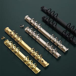 Puede incluir: Cinco anillos de encuadernación metálicos de diferentes tamaños y acabados. Los anillos son plateados, dorados y negros. Los anillos están dispuestos en una fila de izquierda a derecha, con el anillo más grande a la izquierda y el anillo más pequeño a la derecha.