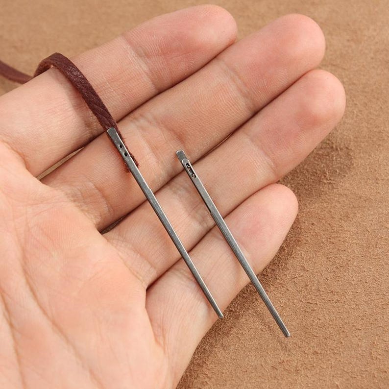 May include: Two silver leather sewing needles with a small eye at the top. The needles are held in a hand against a brown background.