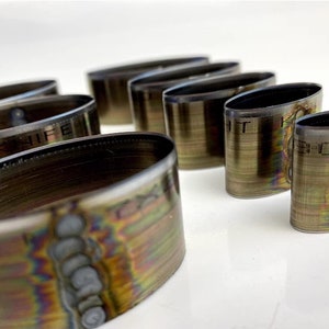 Op de afbeelding: Een set van acht ovale metalen ringen met een regenboogkleurige, iriserende afwerking. De ringen zijn van verschillende grootte en een van de ringen heeft de tekst "T KNIFE" gegraveerd.