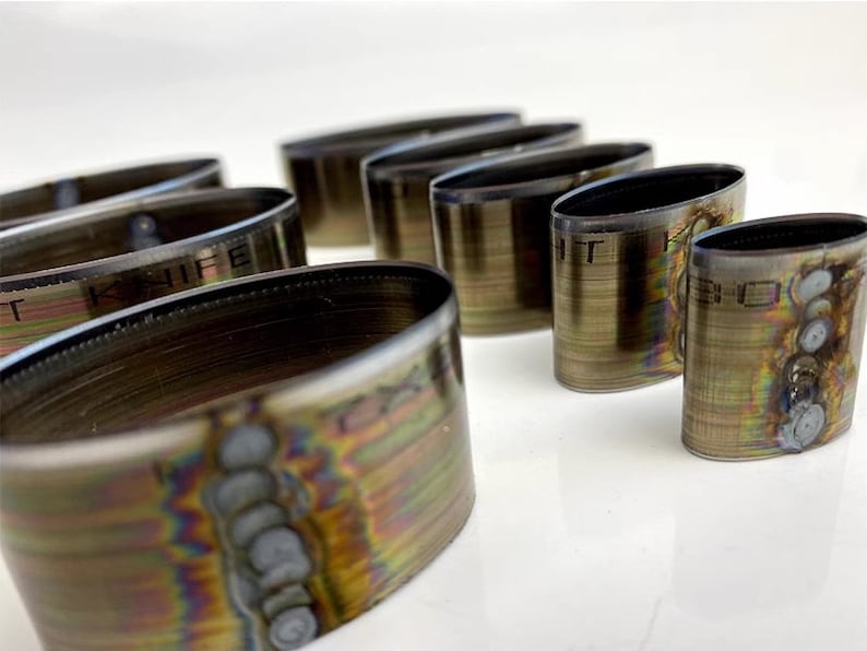 Op de afbeelding: Een set van acht ovale metalen ringen met een regenboogkleurige, iriserende afwerking. De ringen zijn van verschillende grootte en een van de ringen heeft de tekst "T KNIFE" gegraveerd.
