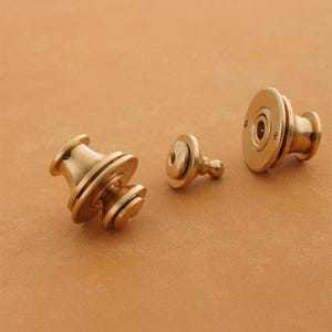 Può includere: Tre chiusure in metallo color ottone con una base rotonda e piatta e un perno cilindrico. Le chiusure sono di dimensioni diverse e hanno un design decorativo e testurizzato.