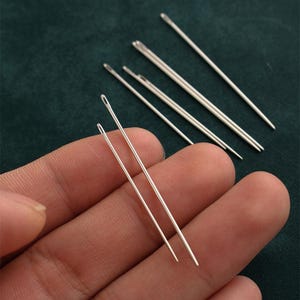 Könnte beinhalten: Nahaufnahme einer Reihe silberner Nähnadeln. Die Nadeln sind spitz und dünn und in Fächerform angeordnet. Die Nadeln werden in einer Hand gehalten, und der Hintergrund ist ein grüner Stoff.