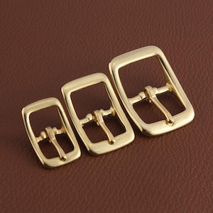 May include: Three gold-toned metal belt buckles of different sizes. The buckles are rectangular with a smooth, polished finish. Each buckle has a center bar with two holes for attaching a belt.