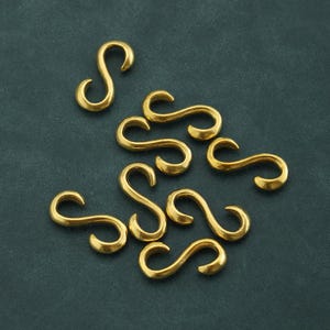 Puede incluir: Un conjunto de diez ganchos en forma de S de metal de color dorado. Los ganchos son todos del mismo tamaño y forma, y están dispuestos en un patrón aleatorio sobre una superficie azul oscuro.