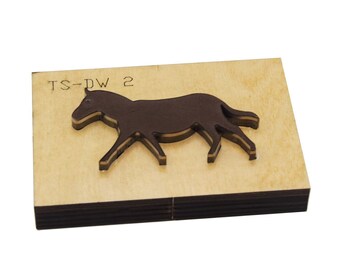 May include: A brown horse-shaped die cut on a wooden base. The die cut is a dark brown color and has a detailed outline. The wooden base has the text "TS-DW 2" etched into the surface. The die cut is likely used for crafting or scrapbooking.