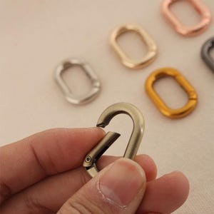 Könnte beinhalten: Ein Satz von fünf Metallringen in verschiedenen Farben und Ausführungen. Die Ringe sind oval geformt und haben eine glatte Oberfläche. Die Ringe werden zum Befestigen an Taschen, Schlüsselanhängern oder anderen Accessoires verwendet.